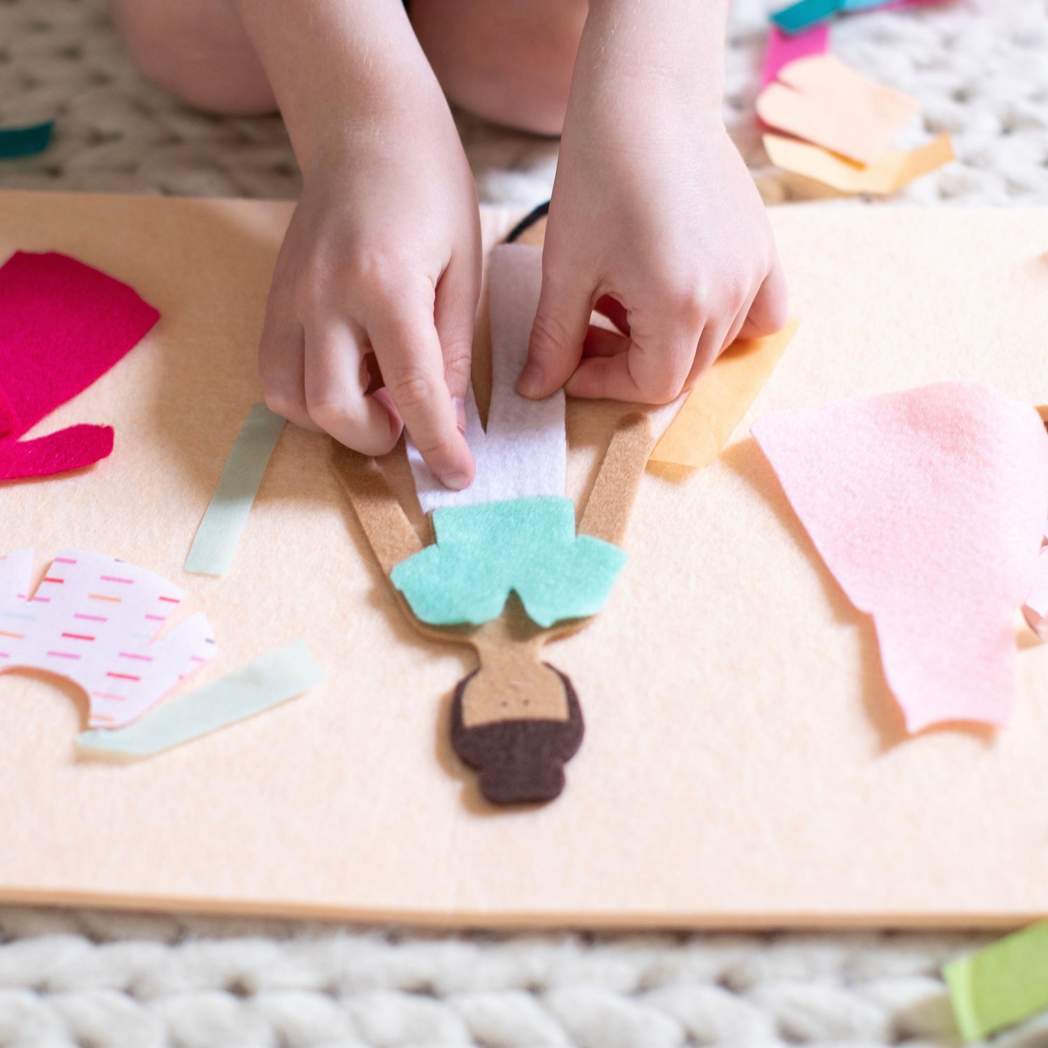 a child dresses a felt fashion doll with turquoise top and white pants. Pink dresses and other clothing pieces are scattered in the background.