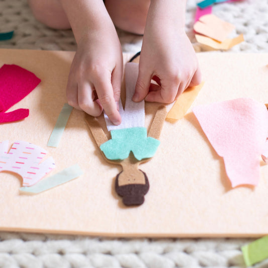a child dresses a felt fashion doll with turquoise top and white pants. Pink dresses and other clothing pieces are scattered in the background.